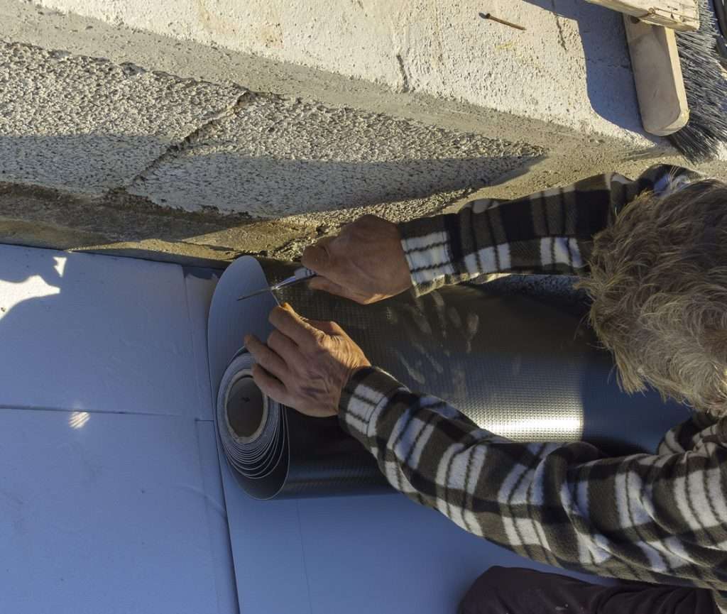 Hands cutting a roll of roofing material on a commercial building, demonstrating installation techniques for flat or low-slope roofs in a construction setting.