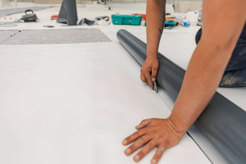 Person installing a single-ply membrane roofing system, using a utility knife on a flat roof surface, surrounded by roofing materials and tools, illustrating commercial roofing installation techniques.