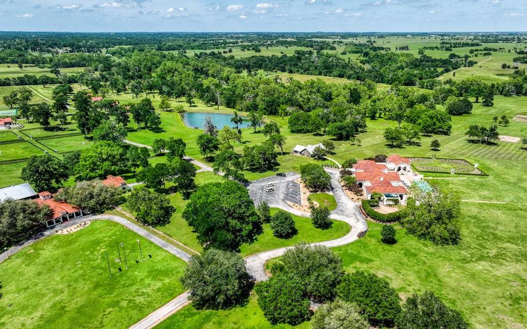 Aerial view of a sprawling ranch in Brenham, Texas, featuring green fields, a pond, and multiple buildings with red roofs, emphasizing the serene landscape ideal for residential or commercial roofing projects.